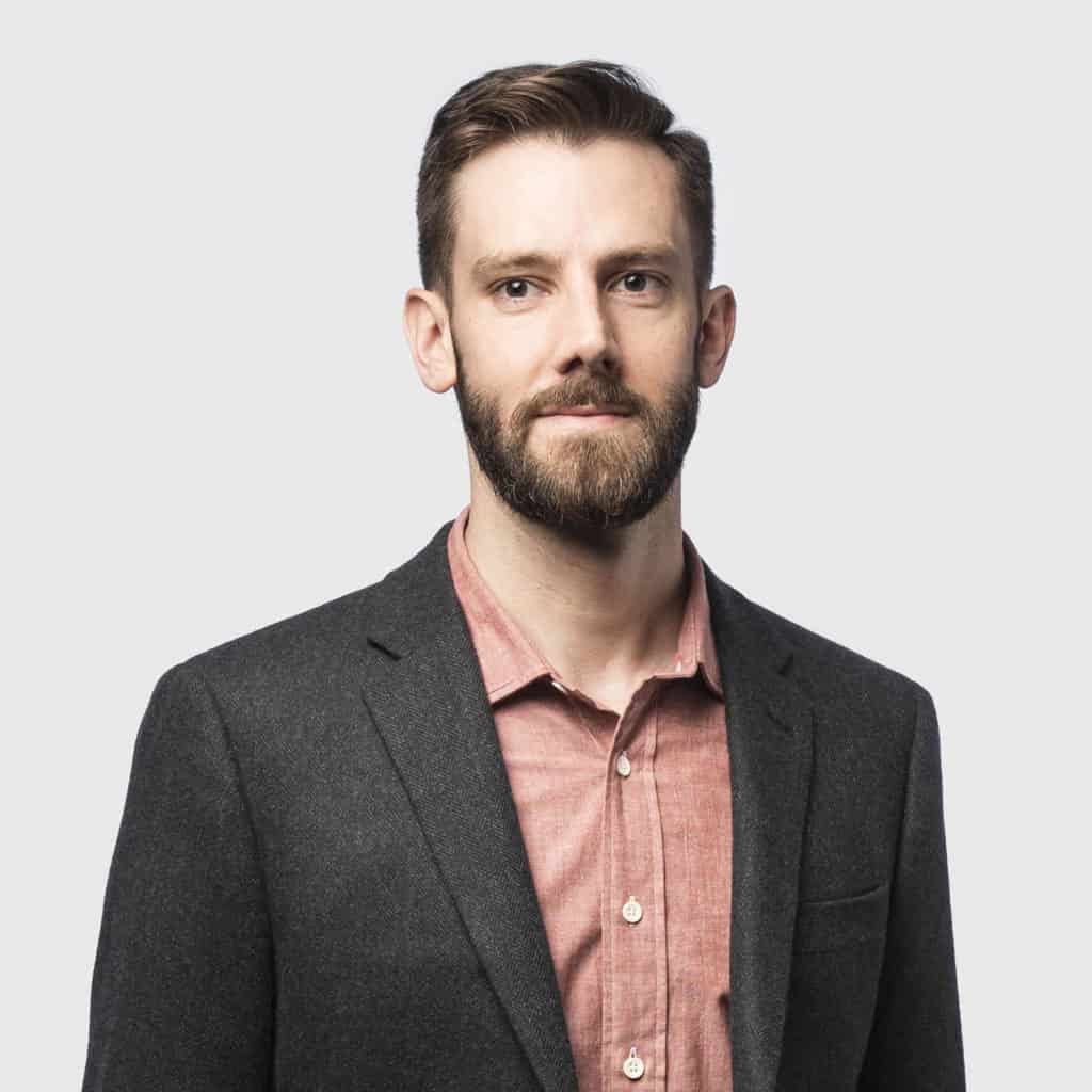 Adult man facing the camera in a dark blazer and button-up shirt, standing against a plain light gray background.