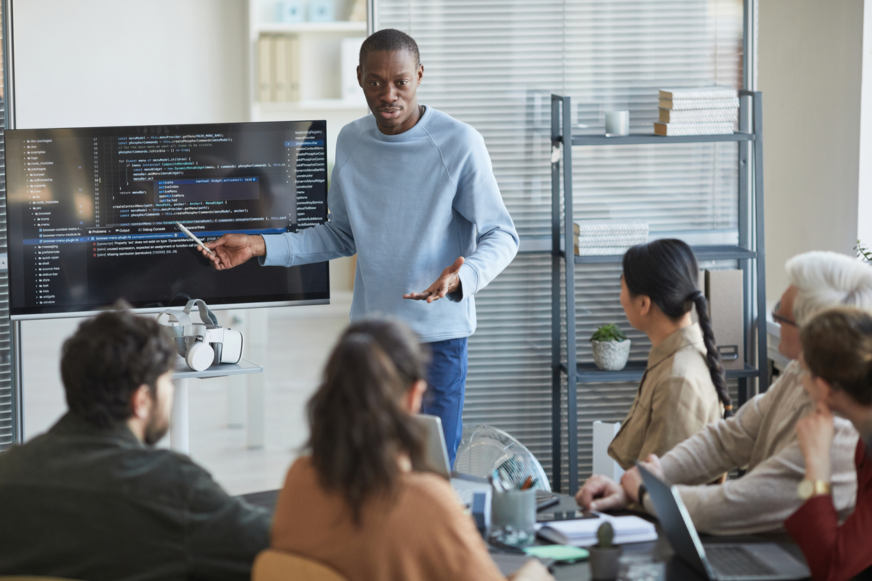 Portrait of emotional African-American man giving presentation in office to IT team and pointing at code on screen, copy space