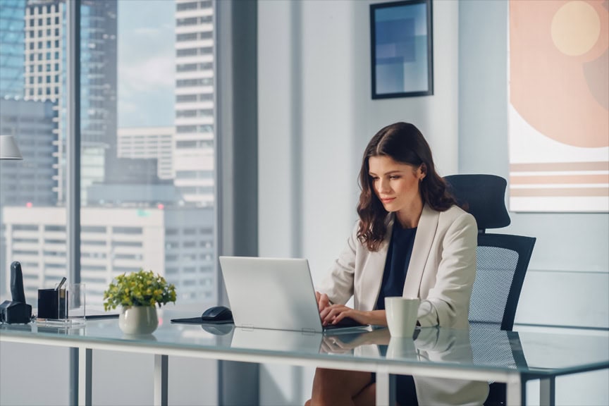 Portrait of Young Successful Caucasian Businesswoman Sitting at Her Desk Working on Laptop Computer in Big City Office. Confident Digital entrepreneur Working on Creative e-Commerce Startup.