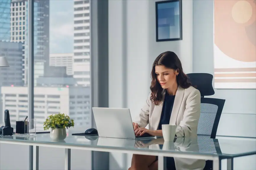 Portrait of Young Successful Caucasian Businesswoman Sitting at Her Desk Working on Laptop Computer in Big City Office. Confident Digital entrepreneur Working on Creative e-Commerce Startup.
