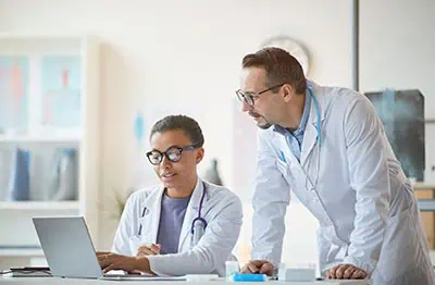 Young nurse sitting at the table and working on laptop and talking to the male doctor in white coat at modern office