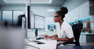 Woman in a white coat works at a desk, viewing two computer monitors and writing on a clipboard in a modern office.